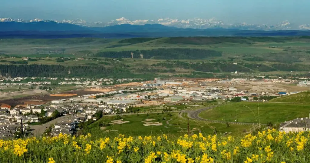 Vast landscape view of a green valley, residential area, and distant snow-capped mountains under clear blue sky.