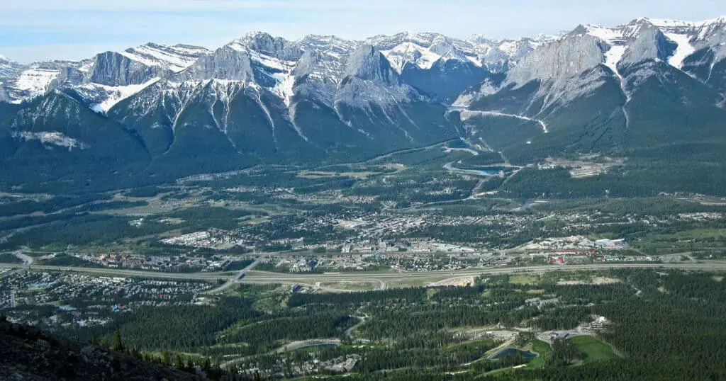 Scenic view of a mountain valley with snow-covered peaks, dense forests, and a winding road, showcasing natural landscapes and outdoor adventure opportunities in the mountainous region.