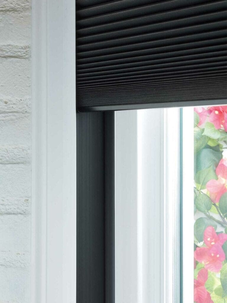 Black vertical window blind on a white frame with a glimpse of colorful flowers outside, offers stylish window covering solutions with premium blinds and drapery services from Blinds West.
